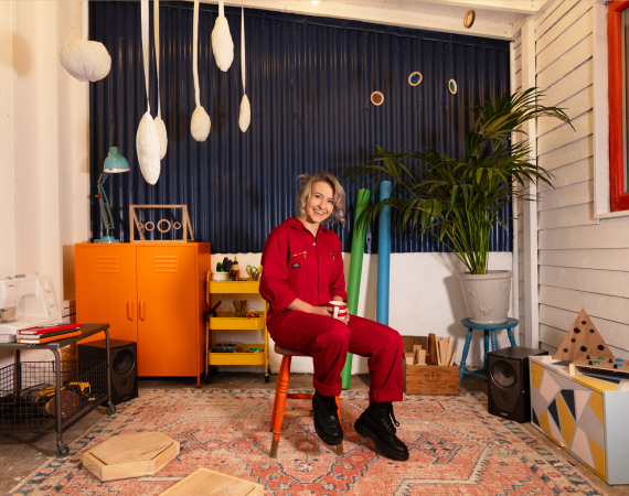 A person with blonde hair wearing a red boilersuit sitting on an orange stall in the middle of a white walled room with a navy corrugated metal panel behind them and pink patterned carpet under their feet. They are surrounded by an orange cupboard, big plant, various tools including a sewing machine and conductive objects they have made hang from the ceiling and sit at the sides of the room 
