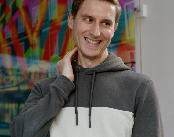 A white man with short, brown hair, wearing a green and white hooded jumper. He is standing up in front of an audience and smiling widely at an audience member