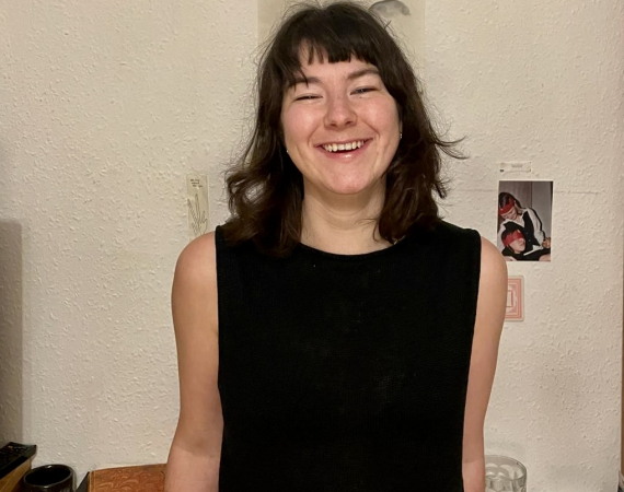 A white woman in her 20s with shoulder length brown hair, wearing a black sleeveless top.