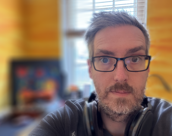 A middle age man with greying hair, glasses, a beard and headphones around his neck