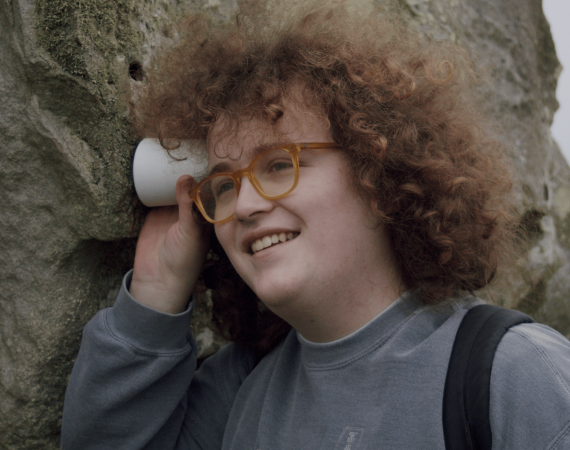 Robin Thorn, a young white person with long curly brown hair is wearing yellow glasses and has their head pressed against a small white cylindrical device that is touching an avebury stone.