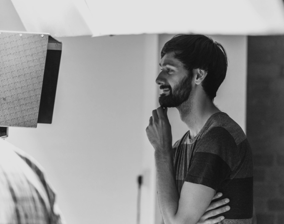 Reuben Armstrong, a white man with short dark hair and a beard, smiling looking at a Director's monitor whilst working on set.