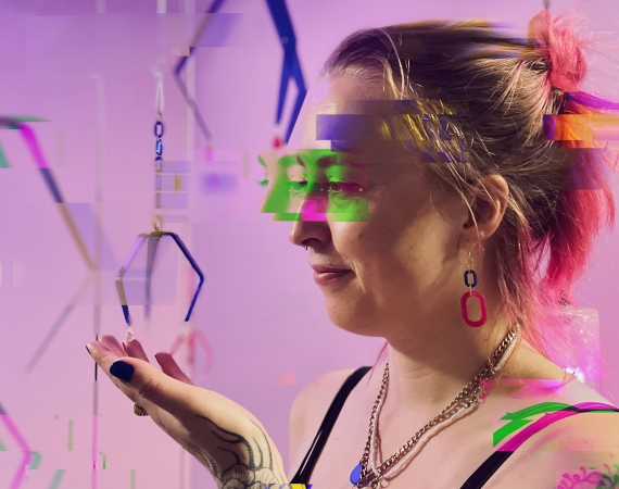 A glitched photo of Suzi Bratt, white female, mid thirties with pink hair and an arm of floral tattoos stands amidst a hanging sculpture of hands and metal arcade grabbers.
