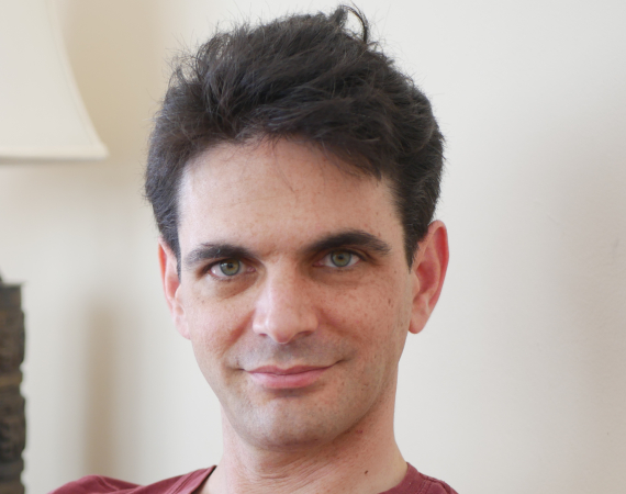 A white man with medium-short black hair, dark eyebrows and freckles. He is wearing a wine-red t-shirt. He is in front of a light background