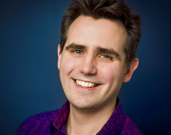 White male with short brown hair, smiling in a purple shirt on a blue background
