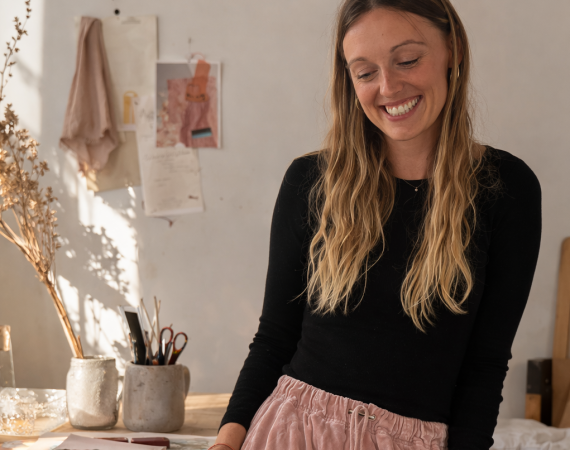 A image of Nina Redman in her Fashion Studio in Bristol, natural light coming into the windows and designs of clothing and samples of fabric on the table in front of her.