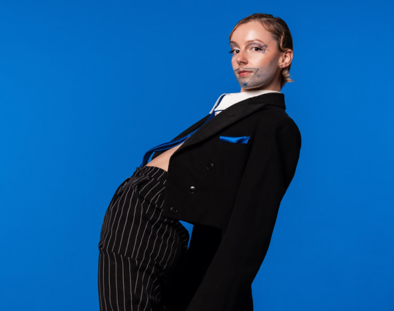 Image is of Joss leaning back wearing a black suit on a striking blue background