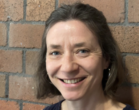 White woman with shortish brown hair and blue eyes, stood in front of a red brick wall