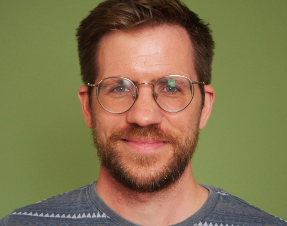 A white man in his mid thirties, with a beard and glasses. He is smiling and standing in front of a green background.