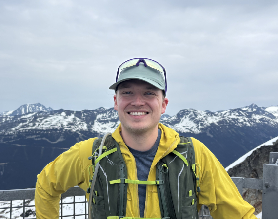 Smiling person wearing a bright yellow jacket and running vest standing in front of a view over a snowy mountain range.