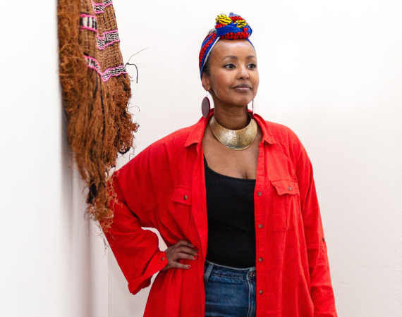 A Somali Woman in a red shirt and blue, yellow and red headwrap next to a grass weaving looking to the distance. 