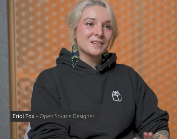 An image of Eriol who is a femme presenting nonbinary person. They are a white person with tattoos on their left arm and wear a black Hoodie with a Penpot logo, have blonde hair and wear Kenyan beaded earrings that are black, blue and yellow