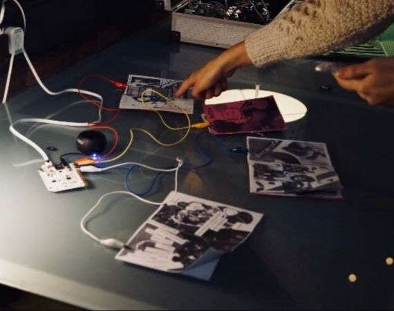 Someone presses a finger to pocket-size zines spread out on a table and wired up to a conductor.