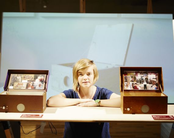 Chloe a white queer woman in her 30s is next to two music memory boxes, her project for people living with dementia.
