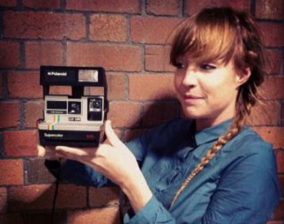 A woman with red hair, wearing a blue shirt and holding a polaroid camera.