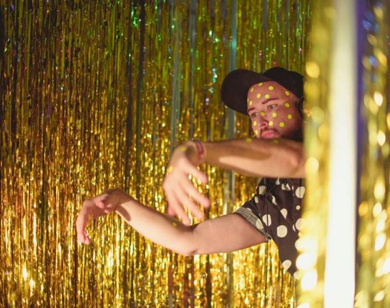 A man stands infront of a gold glitter curtain, wearing a black baseball cap and a black shirt with white spots. 