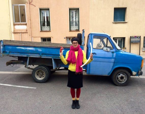 An image of Becca wearing a pink scarf and yellow jumper by a blue van with a cute dog in the drivers seat