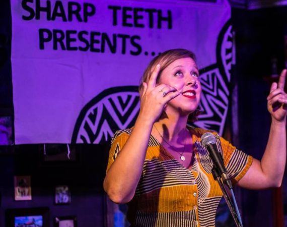 Stephanie points joyfully at the sky as she performs onstage. There is a banner behind her that reads "Sharp Teeth Theatre" and the picture is a delicious mix of rich purples from the light, and golden yellow in Stephanie's outfit. 