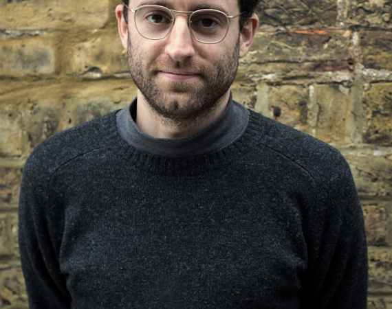 A man with dark hair and silver glasses stands in front of a brick wall. the man is wearing a woollen jumper and is smiling.