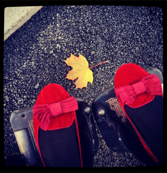 A view of someone looking down at their feet. They are wearing red flat shows with bows on the front, with their feet on two pedals. Between their feet is a yellow, orange toned autumn leaf, 