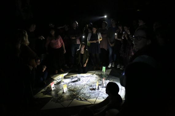 Image of a group of people sat around the edge of a lit up circular installation space on the floor, the circular space is built up of triangles, which resemble a map, and there are little illustrated stands dotted around the space