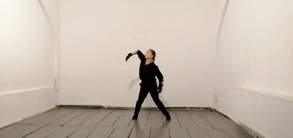 A dancer, dressed all in black, moves around a white brick room. The floor is grey and shiny